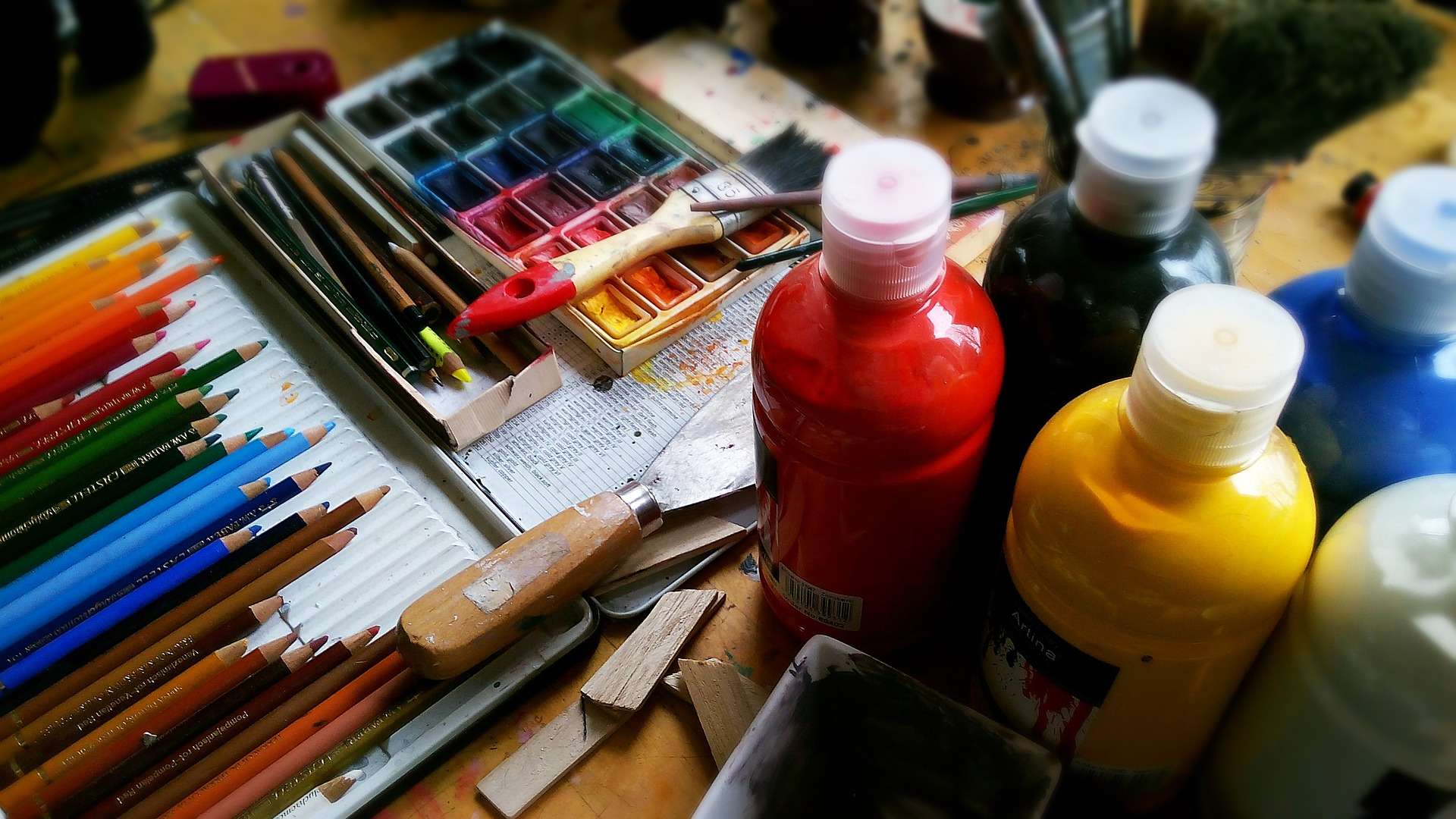 Bottles of paint, coloured pencils and paintbrushes resting on a table.