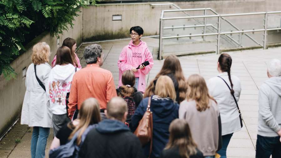 A person in a pink hoodie speaks to a group of people outdoors, near a concrete wall and metal railings.